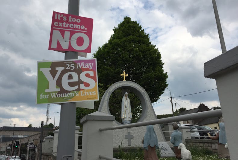 Two signs hang on a lamppost before a Christian statue. One says 